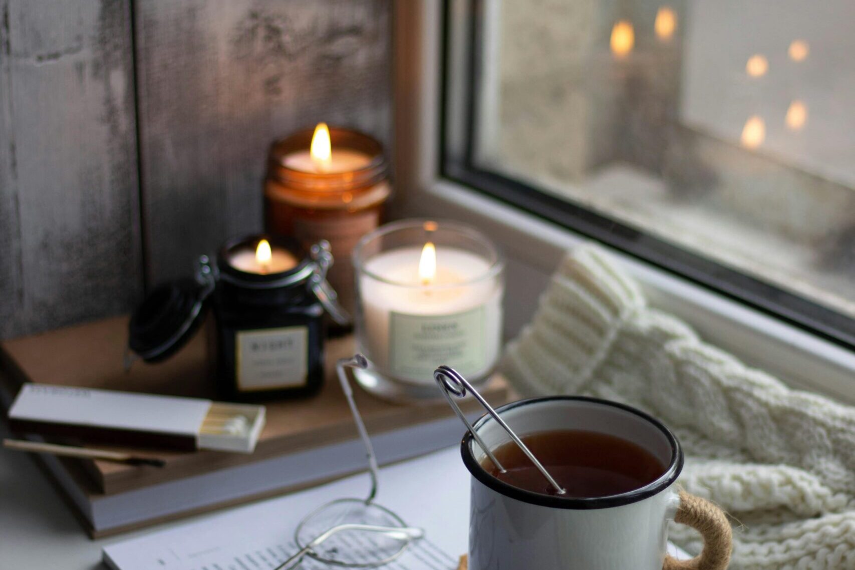 Rincón acogedor junto a la ventana con velas encendidas, un libro, gafas y una taza de té caliente, evocando privacidad e intimidad.