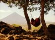 Persona descansando en una hamaca entre árboles con vista a un volcán, disfrutando de la naturaleza y la tranquilidad.