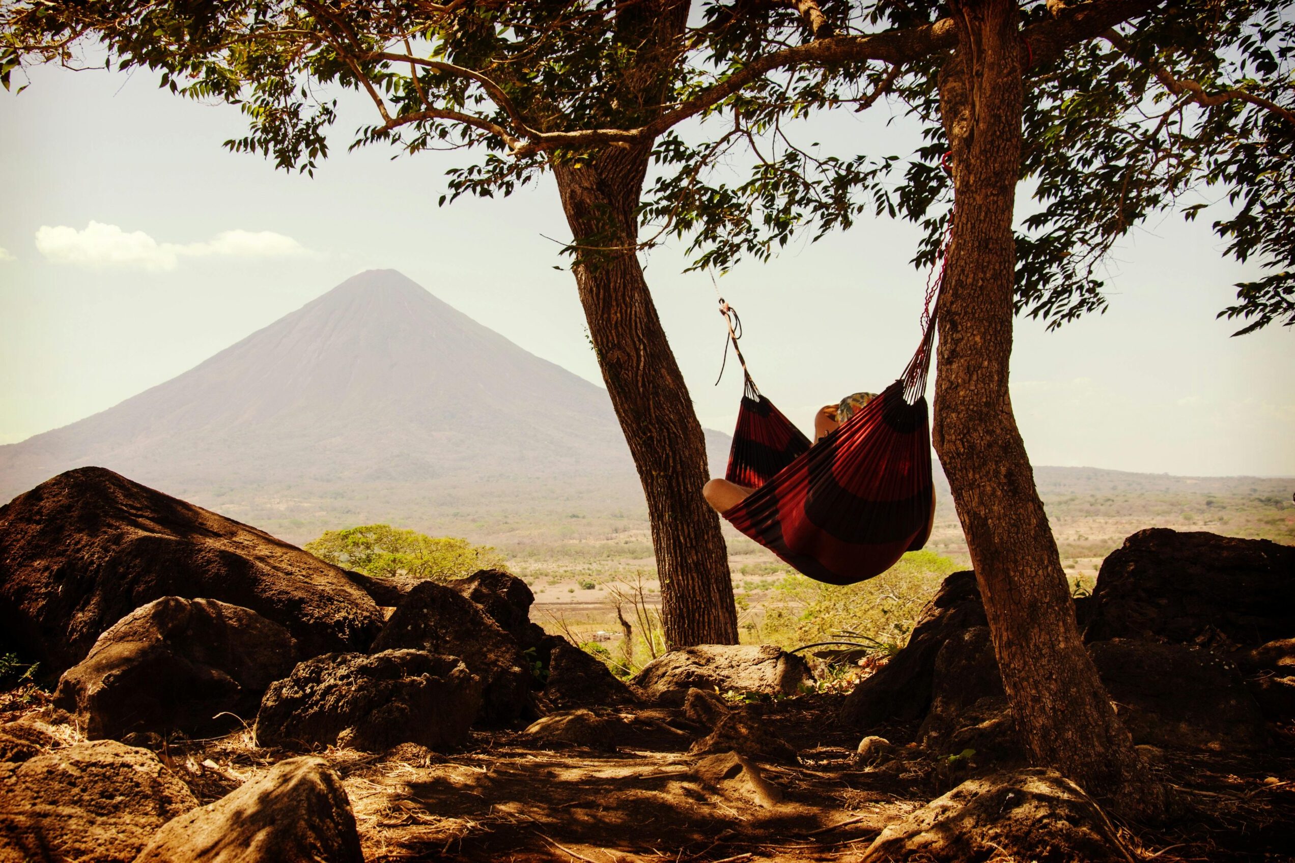 Persona descansando en una hamaca entre árboles con vista a un volcán, disfrutando de la naturaleza y la tranquilidad.
