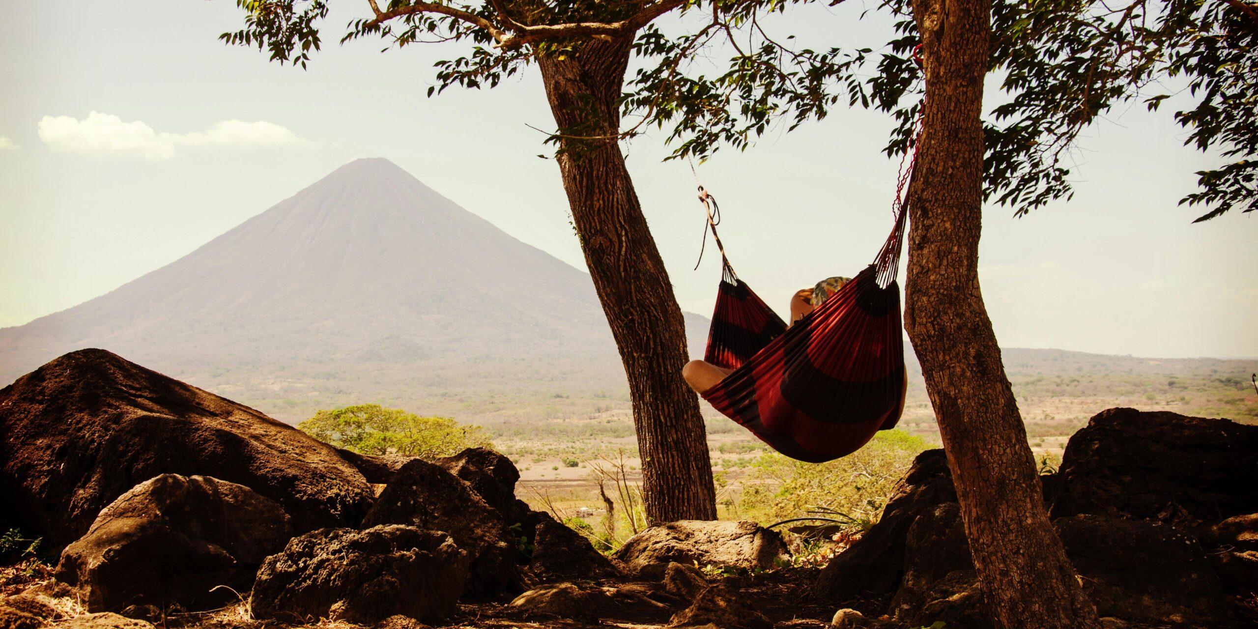 Persona descansando en una hamaca entre árboles con vista a un volcán, disfrutando de la naturaleza y la tranquilidad.