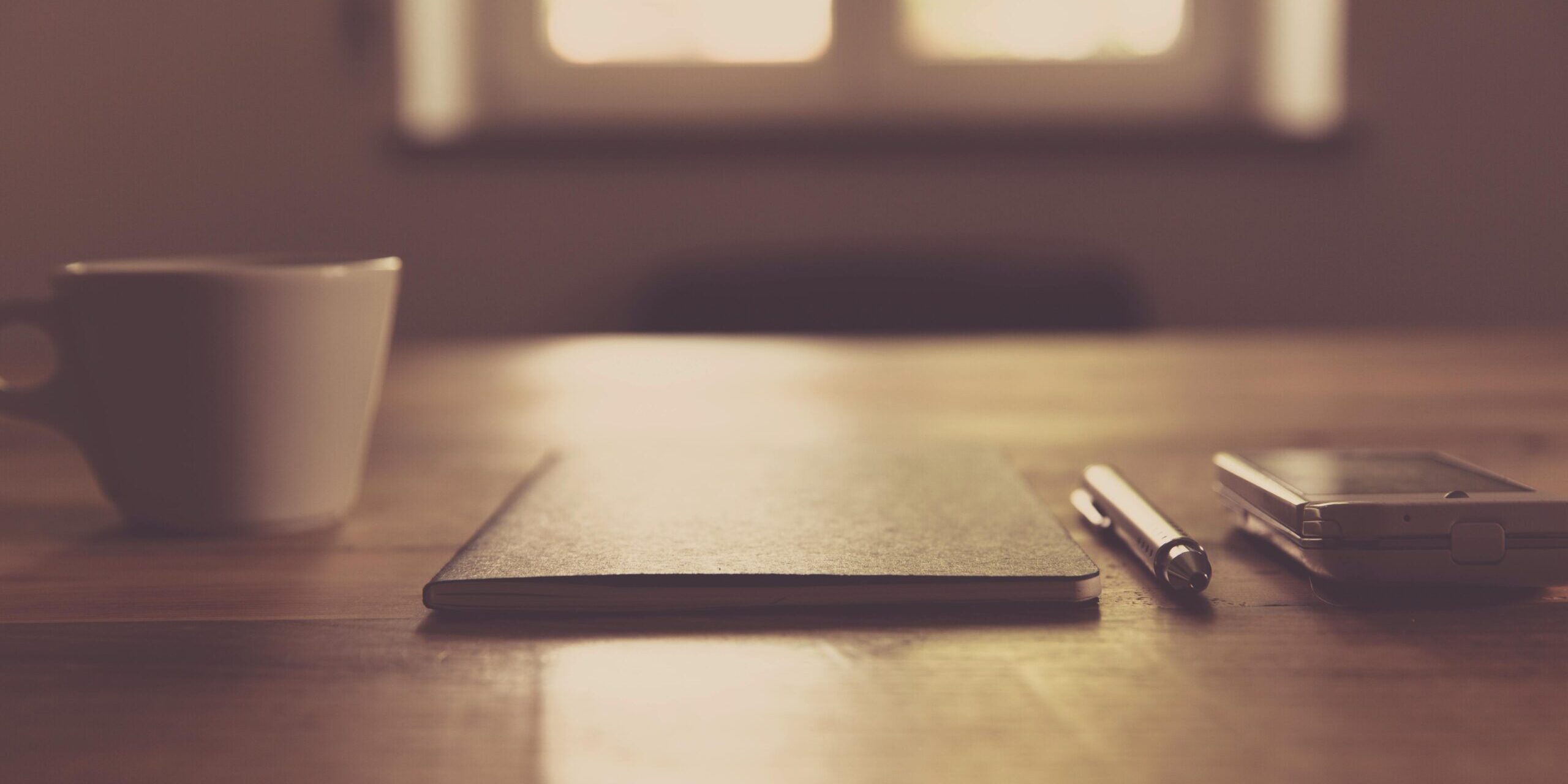 Mesa minimalista con una taza de café, un cuaderno, bolígrafo y un smartphone junto a una ventana, representando calma y enfoque.