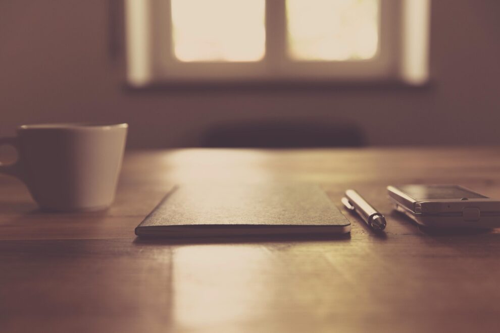 Mesa minimalista con una taza de café, un cuaderno, bolígrafo y un smartphone junto a una ventana, representando calma y enfoque.